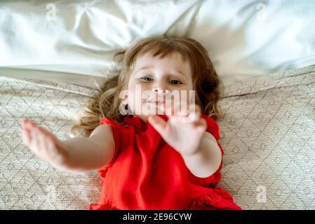 Baby Mädchen hält ihre Hände mit einer Anfrage Top View. Schlechte Laune, weint und fragt, besteht und fordert. Liegend auf dem Bett. Bittet um Hilfe, Stockfoto