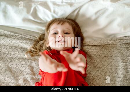 Baby Mädchen hält ihre Hände mit einer Bitte. Schlechte Laune, weint und fragt, besteht und fordert. Liegend auf dem Bett. Bittet um Hilfe, Top View Stockfoto