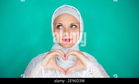 Schöne junge muslimische Frau in weiß gibt ein Symbol des Herzens mit ihren Händen. Konzept der Liebe, Valentinstag. Stockfoto