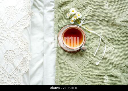 tasse natürlichen Kräutertee aus Minze und Zitronenmelisse im Bett, am Morgen Nahaufnahme. Gemütliche Atmosphäre. Durchbrochene Spitze, weiße Baumwolldecke, Sommer-Gänseblümchen-Blumen Stockfoto