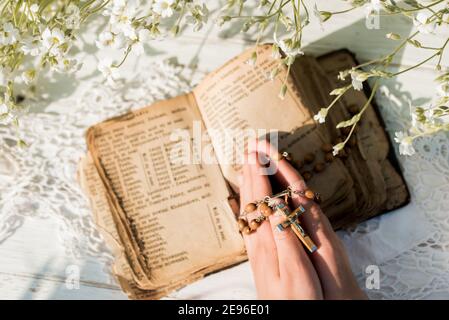 Hände gefaltet im Gebet über alte Bibel. Hölzerner Hintergrund.Hände und Rosenkranz, Gebet, altes Buch mit gelben Seiten. Weiße Blumen auf einem Hintergrund. In Stockfoto