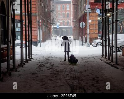New York, New York, USA. Februar 2021. Eine Mutter zieht ihren Sohn auf einem Schlitten durch die Staple Street in Tribeca. Diese ikonische NYC Straße mit ihrer Skybridge, die jetzt von Gerüsten umschlossen ist, wurde in unzähligen Filmen und tv-Shows gezeigt. Quelle: Milo Hess/ZUMA Wire/Alamy Live News Stockfoto