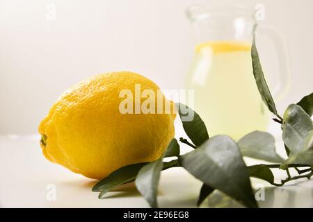 Zitrone und Blätter auf weißem Tisch und Krug mit Wasser mit Zitrone im Hintergrund. Vorderansicht. Horizontale Zusammensetzung. Stockfoto