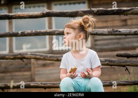 Outdoor-Porträt eines Mädchens auf dem Gras in der Nähe des Zauns sitzen.Sommer im Dorf. Schöne Baby-Mädchen auf einer Holzbank.Ökologie und glücklich Stockfoto