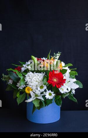 Helles Bouquet von Gerberas, Chrysanthemen, Lilien, grünen Blättern in einer Geschenkbox auf dunklem Hintergrund. Eine Karte mit floralem Hintergrund für jeden Anlass. Stockfoto