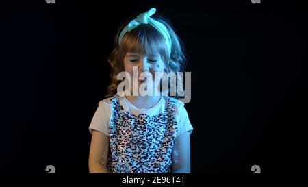 Portrait von trendy stilvolle Mädchen zeigt Zunge, um zu täuschen. Elegantes Kind Kind in Neonbeleuchtung posiert bei Disco Party Cyberpunk Studio. Futuristische Hipster Teenager Kinder machen Gesichter Stockfoto