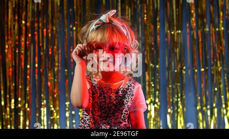 Portrait von trendy stilvolle Mädchen mit Blick auf die Kamera, lächelnd. Nettes Kind Kind in roten Neonbeleuchtung posiert in Disco Cyberpunk Club. Futuristische Hipster Teenager Kinder machen Gesichter, um zu täuschen Stockfoto