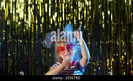 Stilvolle Teenager Kind Mädchen auf hinterleuchteten blauen Neon-Lichter stehen und zeigt auf leeren Raum, zustimmen Zeichen, um zu täuschen. Kind Blick auf die Kamera im Nachtclub. Produkt, Logo oder Text präsentieren. Kopierbereich Stockfoto