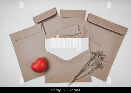 Leeres weißes Papier wird auf den offenen braunen Papierumschlag mit rotem Herz und Limonium-Trockenblume auf weißem Hintergrund gelegt. Valentinstag-Konzept. Stockfoto