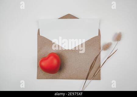 Weißes Blankopapier wird auf einen offenen braunen Papierumschlag mit rotem Herz mit Hasenschwanz trockener Blume auf weißem Hintergrund gelegt. Valentinstag-Konzept. Stockfoto