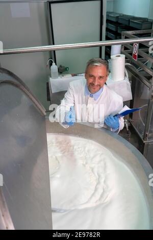 Lebensmittelingenieur in weißer Uniform und blauen Handschuhen mit Blick auf die Kamera. Pasteurisierung der Milch in der Verarbeitungsanlage für Milchprodukte. Lebensmittelsicherheit In Molkereianlagen. Stockfoto