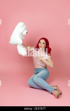 Frau hält großen weißen Teddybär sitzen auf dem Boden und Punkte mit Zeigefinger isoliert auf rosa Hintergrund Stockfoto