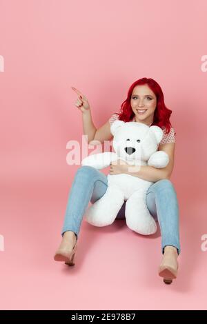 Frau in Jeans mit roten Haaren hält ein großes Weiß teddybär und zeigt mit ihrem Zeigefinger auf leer Leerzeichen für Text Stockfoto