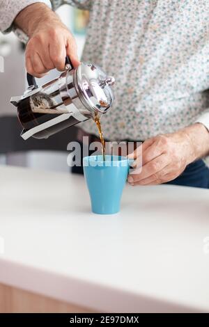 Älterer Mann, der beim Frühstück in der Küche Kaffee mit einer französischen Presse zubereitet und in eine blaue Tasse gießt. Alte Person am Morgen genießen frische braune Café Espresso Tasse Koffein, Filter entspannen Erfrischung Stockfoto