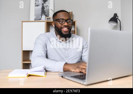 Glücklich und aufgeregt afroamerikanischen Unternehmer in Brillen im Gespräch mit Geschäftspartnern auf dem Video-Anruf und tippen, Notizen machen, und aufmerksam Stockfoto