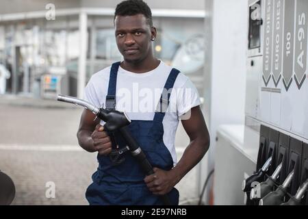 Hübscher junger afrikanischer Kerl, Tankstellenarbeiter, trägt weißes T-Shirt und blaue Overalls und hält Füllpistole. Nahaufnahme der Vorderansicht Stockfoto