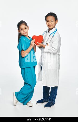 Gruppe von multiethnischen Kindern tragen medizinische Uniformen, halten Kinder Form des Herzens in den Händen. Zukünftige Herzärzte Stockfoto