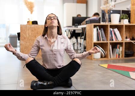 Ziemlich entmutigende hispanische Frau meditiert auf dem Boden im Büro sitzen. Stockfoto
