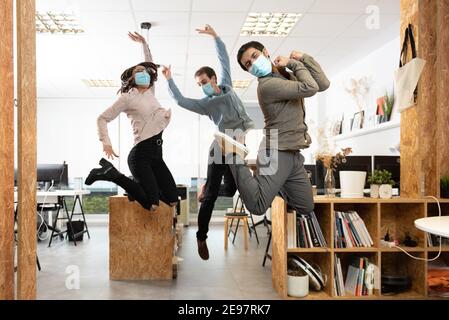 Glückliche Mitarbeiter tragen Schutzmasken springen und feiern am Arbeitsplatz. Arbeiten im Büro während des Coronavirus-Pandemiekonzepts. Stockfoto