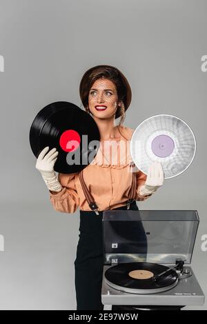 Glückliche Frau in Vintage-Kleidung hält Vinyl-Discs in der Nähe von Rekord Player auf Grau isoliert Stockfoto