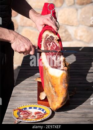 Mann schneidet eine Scheibe getrockneten spanischen Jamon serrano auf Holztisch. Stockfoto