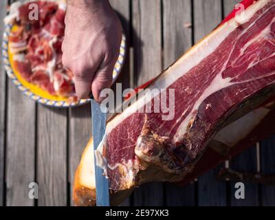 Mann schneidet eine Scheibe spanischen getrockneten Schinken und Teller mit den Scheiben auf dem Hintergrund. Stockfoto