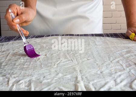 Frau und kleines Mädchen schmieren rohen Teig auf die gelegt Tisch mit einem Pinsel Stockfoto