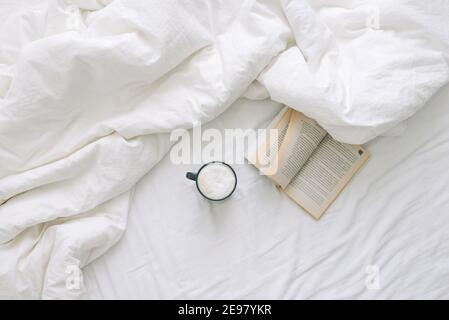 Eine Tasse Kaffee und ein offenes Buch liegen auf einem weißen, offenen Bett. Draufsicht Stockfoto