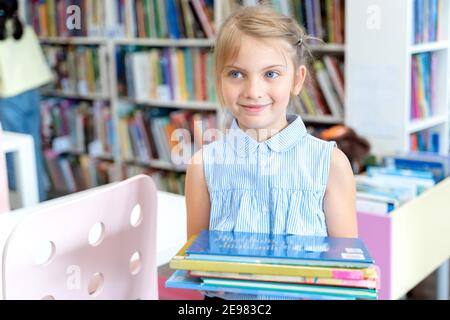 Kleines Mädchen hält Stapel von Büchern mit Märchen in der Kinderbibliothek. Kinderzimmer mit Sonderlesung. Regale mit hellen interessanten, lehrreichen Büchern Stockfoto