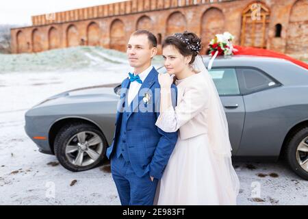 Die Braut und der Bräutigam in der Nähe des Autos, umarmen und küssen ihre Liebsten bei der Hochzeit, die Familie beim Winterfotoschießen mit der Hochzeitskortege Stockfoto