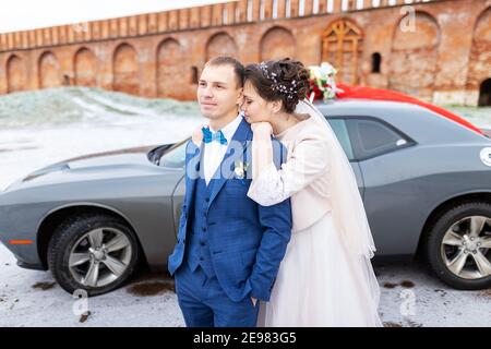 Die Braut und der Bräutigam in der Nähe des Autos, umarmen und küssen ihre Liebsten bei der Hochzeit, die Familie beim Winterfotoschießen mit der Hochzeitskortege Stockfoto
