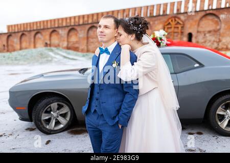 Die Braut und der Bräutigam in der Nähe des Autos, umarmen und küssen ihre Liebsten bei der Hochzeit, die Familie beim Winterfotoschießen mit der Hochzeitskortege Stockfoto