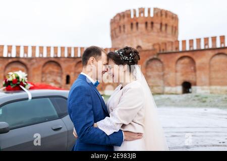 Die Braut und der Bräutigam in der Nähe des Autos, umarmen und küssen ihre Liebsten bei der Hochzeit, die Familie beim Winterfotoschießen mit der Hochzeitskortege Stockfoto