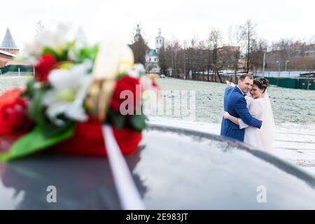 Die Braut und der Bräutigam in der Nähe des Autos, umarmen und küssen ihre Liebsten bei der Hochzeit, die Familie beim Winterfotoschießen mit der Hochzeitskortege Stockfoto