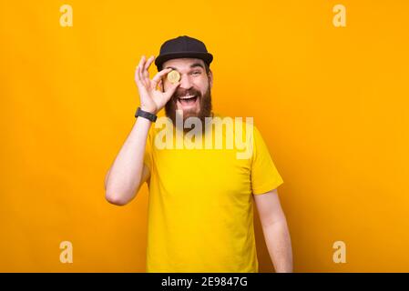 Foto von glücklich lächelnden Mann mit Bart hält Bitcoin über den Augen. Stockfoto