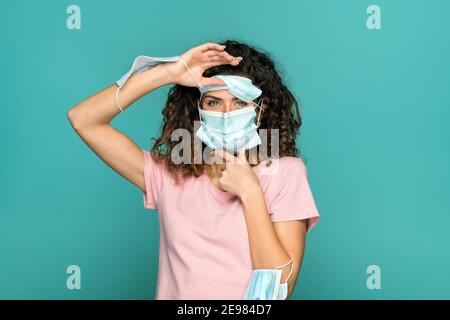 Hübsche Frau, die viele medizinische Masken auf Gesicht und Armen trägt Beim Schutz vor Coronavirus und Blick auf Kamera auf blau Hintergrund im Studio Stockfoto