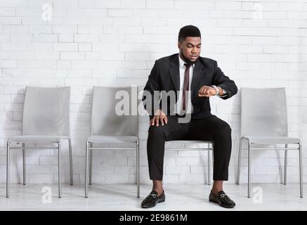 Junger afroamerikanischer Mann in formeller Kleidung, der seine Uhr anschaut, die Zeit überprüft und auf ein Vorstellungsgespräch in der Bürolobby wartet Stockfoto