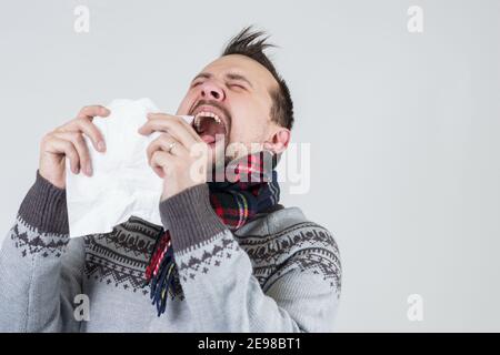 Junge kaukasische kranke Mann trägt Schal und grauen Pullover niesen Mit offenem Mund mit Ausdruck wegen winterlicher Kälte Oder Allergie Stockfoto