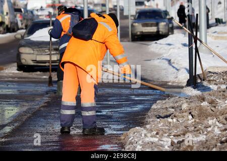 Schneeräumung in der Winterstadt, Straßenreinigung. Zwei kommunale Arbeiter in orangefarbener Uniform mit einer Schaufel räumen schmelzenden Schnee auf einem Bürgersteig Stockfoto