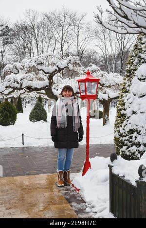 Junge lächelnde Frau, die in der Nähe einer roten Laterne im alten Stil mit weihnachtsdekoration in einem schneebedeckten Winterpark steht. Schneebedeckter Winter Stockfoto