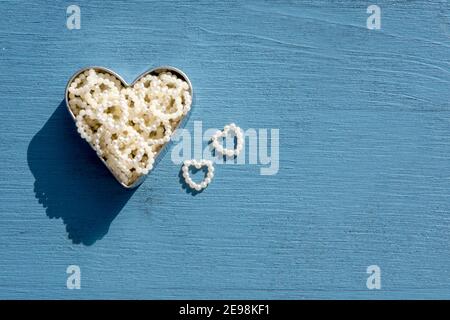 Celebration Love Concept: Draufsicht auf zwei kleine Perlherzen neben einem großen metallischen Herz gefüllt mit vielen kleinen Herzen auf hölzernem blauem Hintergrund Stockfoto