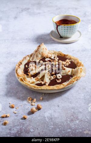 Teller mit gefalteten Crepes (oder Pfannkuchen) mit dunkler Schokoladensauce und gerösteten Haselnüssen bedeckt, Krug mit Schokoladensauce im Hintergrund. Stockfoto