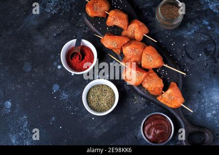 Rohe Hähnchenspieße in Marinade mit Gewürzen auf einem schwarzen Brett. Rohes Hähnchen-Shish-Kebab auf Spieße mit Gewürzen auf dunklem Hintergrund. Stockfoto