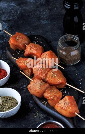 Rohe Hähnchenspieße in Marinade mit Gewürzen auf einem schwarzen Brett. Rohes Hähnchen-Shish-Kebab auf Spieße mit Gewürzen auf dunklem Hintergrund. Stockfoto