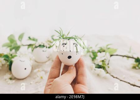 Hand halten osterei mit gezeichnet lustigen Gesicht in Blumenkranz auf Hintergrund von natürlichen Eiern und Blütenblätter auf Leinenstoff in weichem Licht. Frohe Ostern und Stockfoto