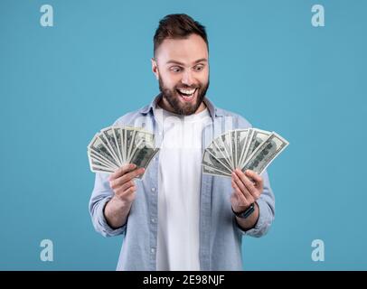 Wohlhabender Kaukasischer Kerl mit zwei Fans auf Geld begeistert über seinen großen Erfolg über blauen Hintergrund. Riesiges Gewinnkonzept Stockfoto