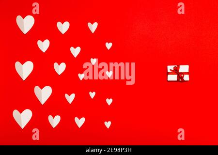 Valentinstag Hintergrund. Weiße Papierherzen und kleine Geschenkschachtel auf rotem Hintergrund in flacher Lay-Optik. Valentinstagskonzept Stockfoto