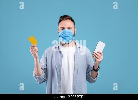 Kontaktloses Einkaufen während der covid Quarantäne. Junger Mann in Maske hält Handy und Kreditkarte, blauer Hintergrund Stockfoto