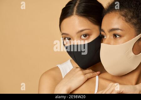 Zwei entzückende junge Frauen, weibliche Freunde tragen schützende Gesichtsmasken, während zusammen posiert auf beigem Hintergrund isoliert. Sicherheit, Pandemiekonzept Stockfoto