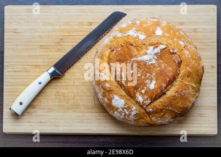 Frisch gebackenes braunes Brot mit herzförmiger Kruste und Ein Brotmesser auf einem Holzteller Stockfoto